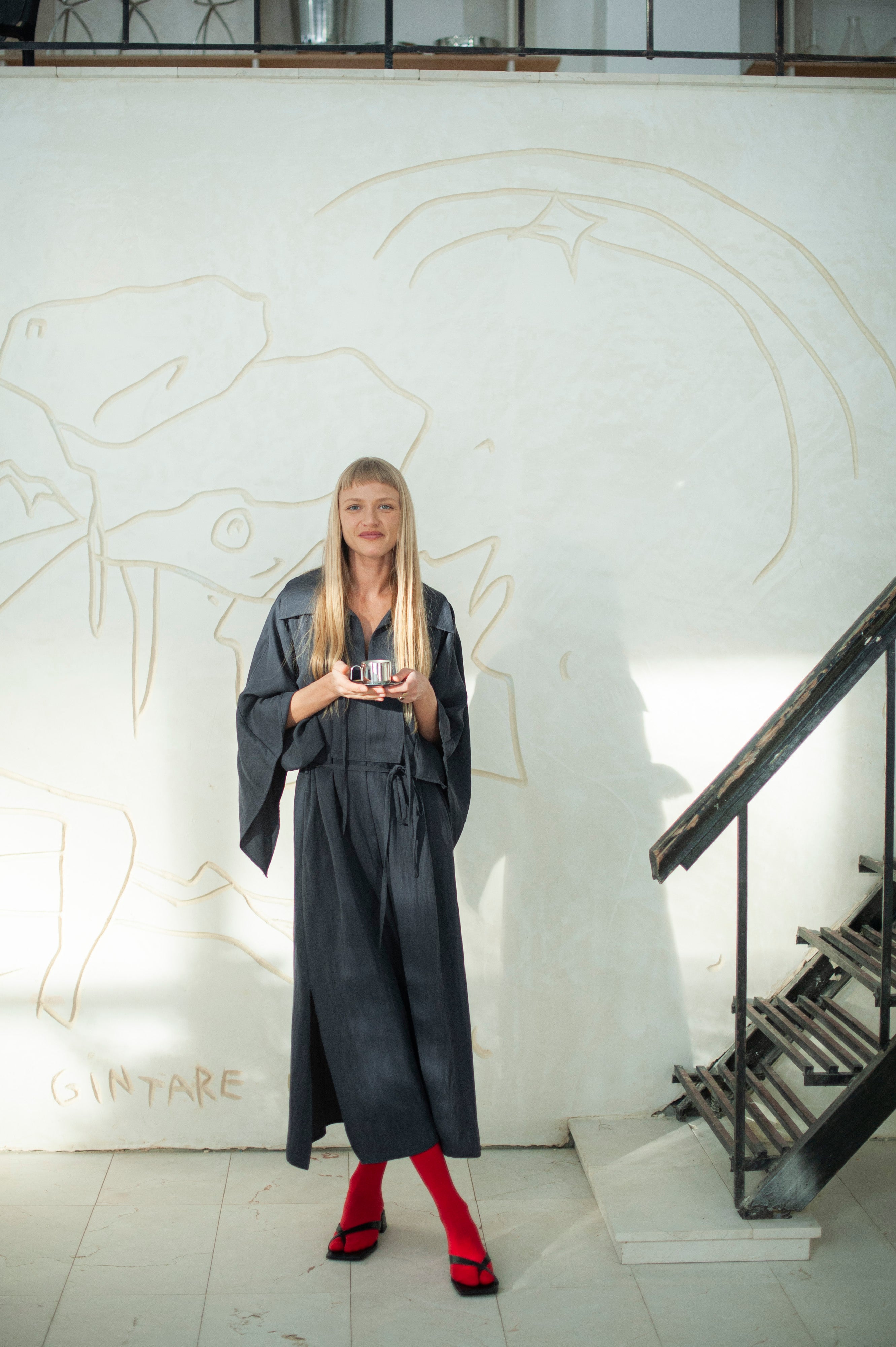 A blonde wearing a long, flowy gray cupro dress with wide kimono-inspired sleeves featuring side slits. The dress has a slightly oversized collar on a subtle V-neckline and a waist tie that gently defines the silhouette.