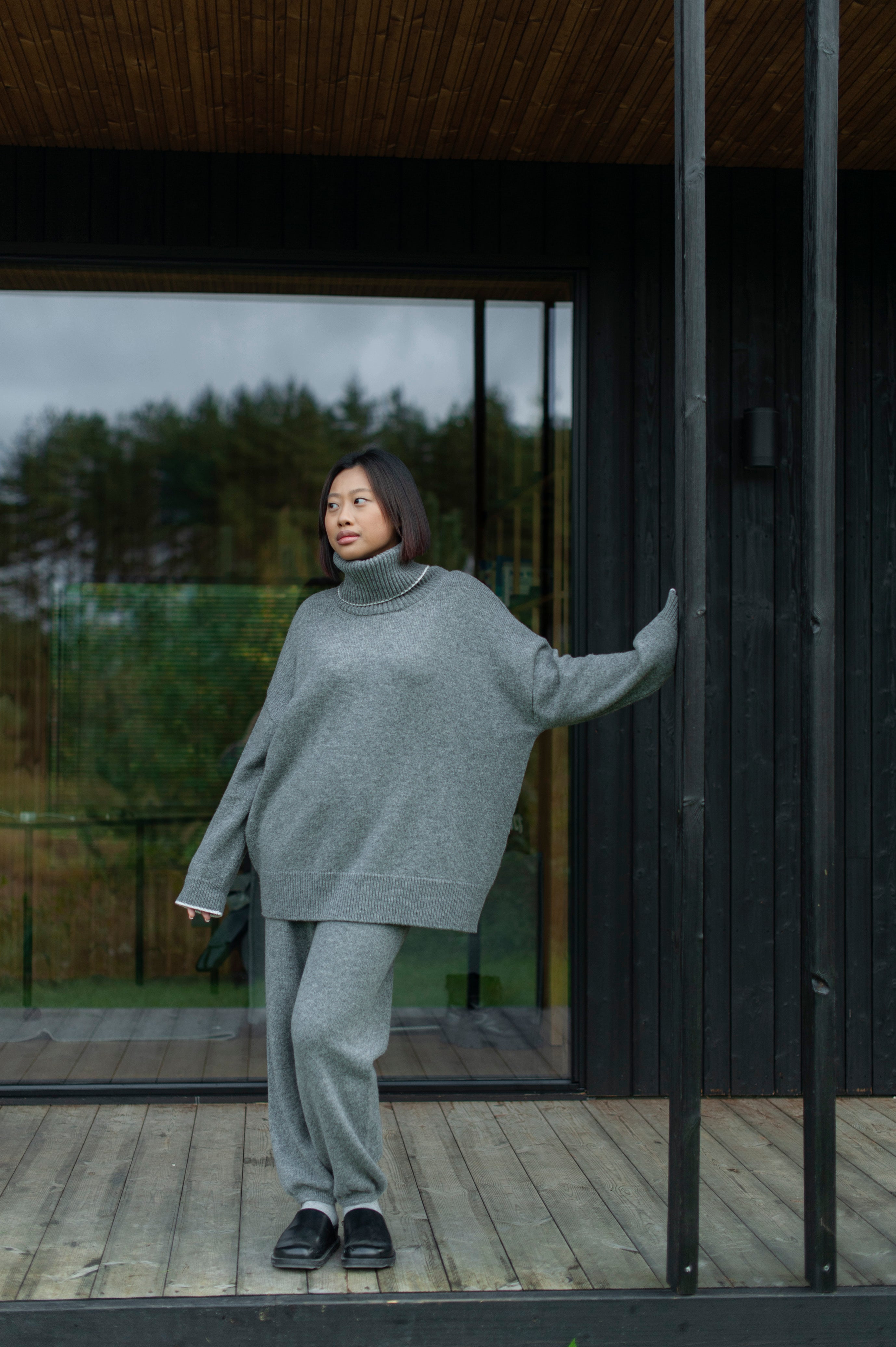 Person wearing a gray sweater and pants standing on a wooden deck with a modern building in the background.