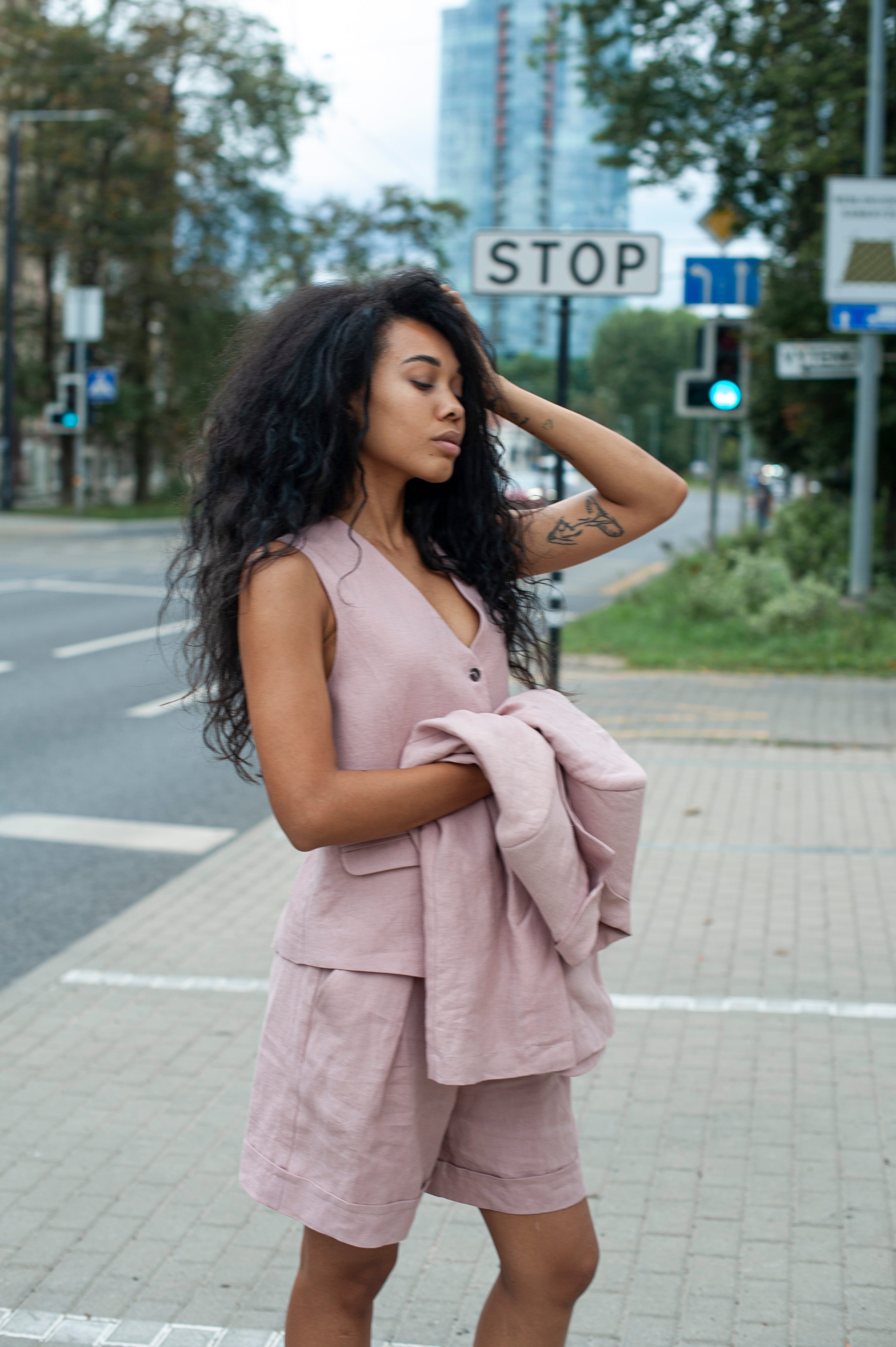 A women's three-piece ensemble featuring a linen vest and pleated shorts. The woman is also holding a matching linen blazer.