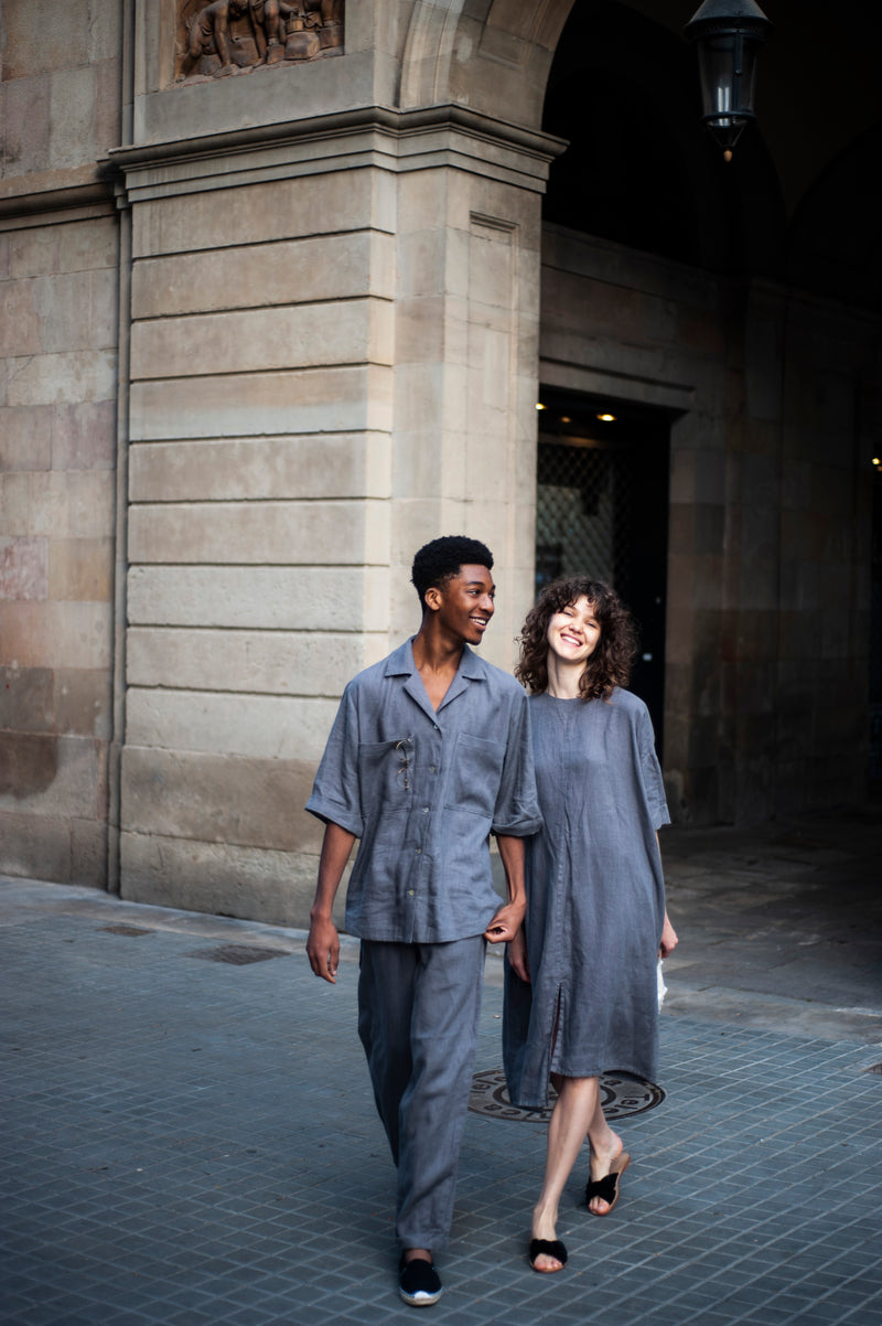 A couple in grey matching linen outfits. The man is in a two-piece linen set featuring a camp collar shirt and pants, and the girl is wearing an asymmetric  oversized dress with elbow-length sleeves