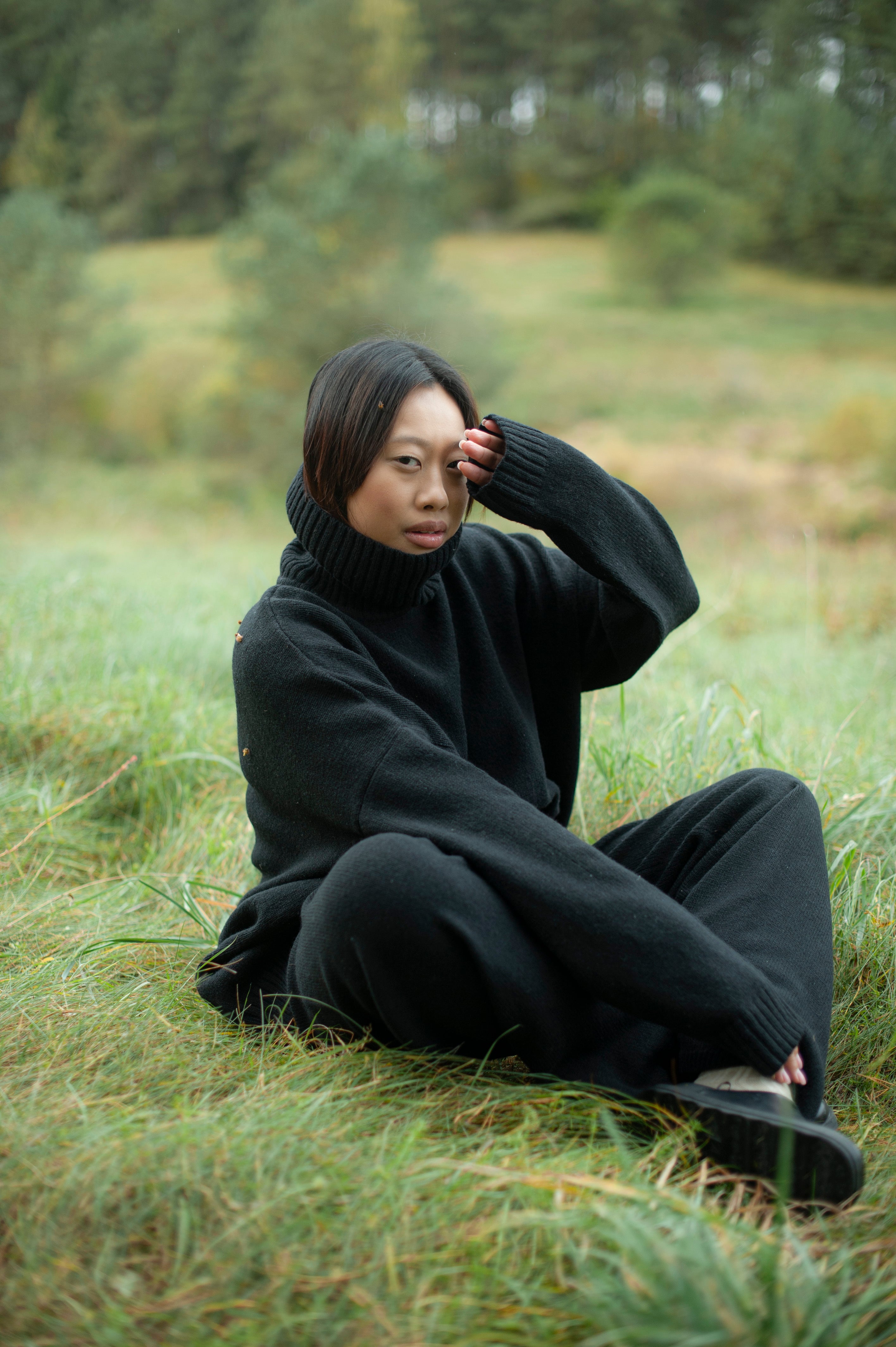 A lady in an overside knit roll-neck sweater, sitting on the grass in the nature. 