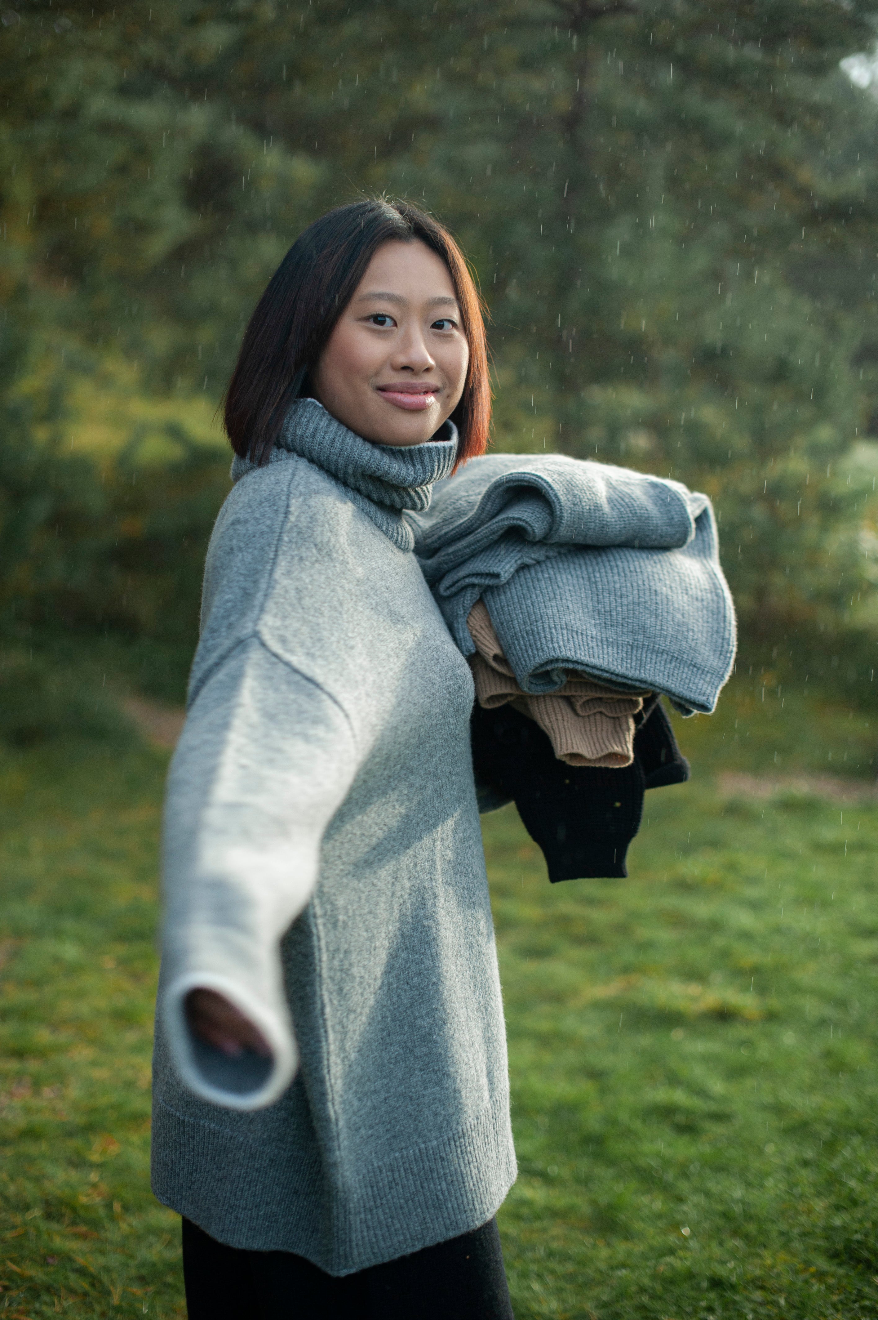 A girl in a grey merino wool jumper smiling in the rain and holding different color sweaters