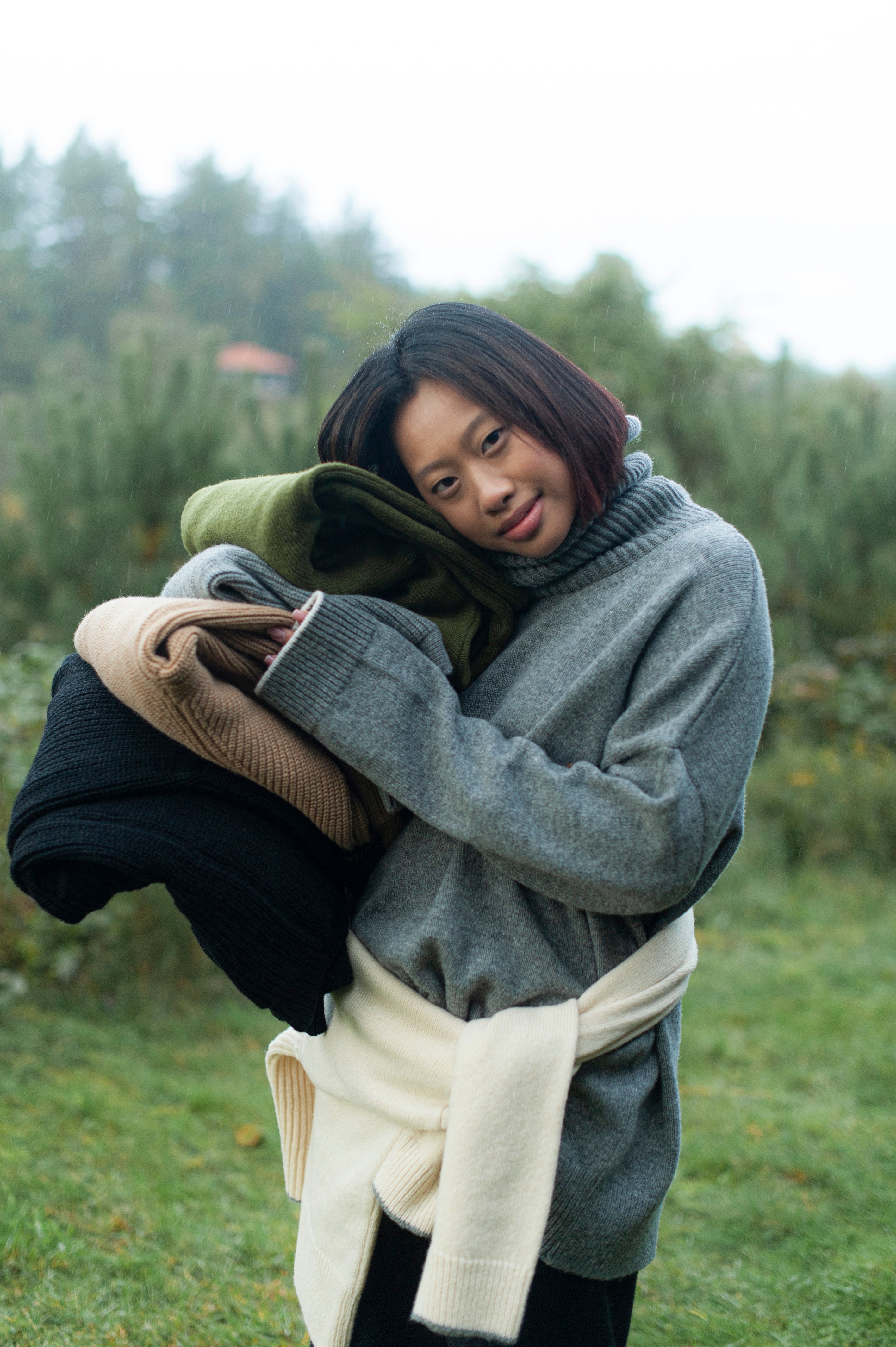 A model in a grey high-neck jumper, holding a pile of women's merino wool knitwear. 